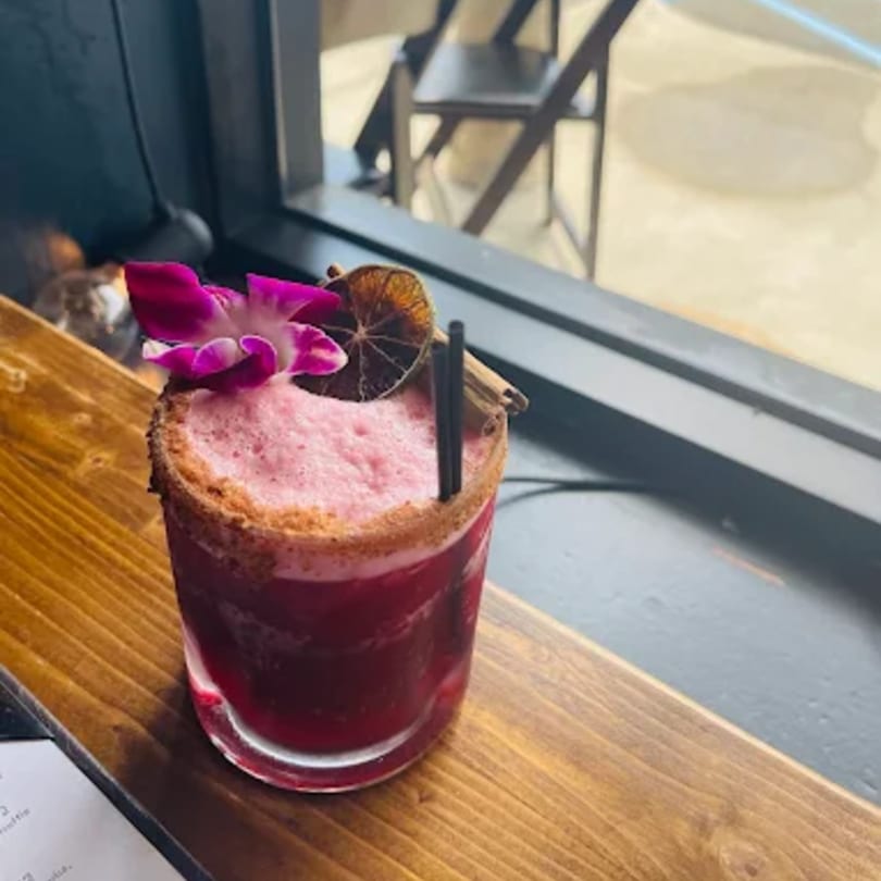 A vibrant red Caribbean cocktail with pink foam, garnished with an orchid, dried orange, and cinnamon sticks.