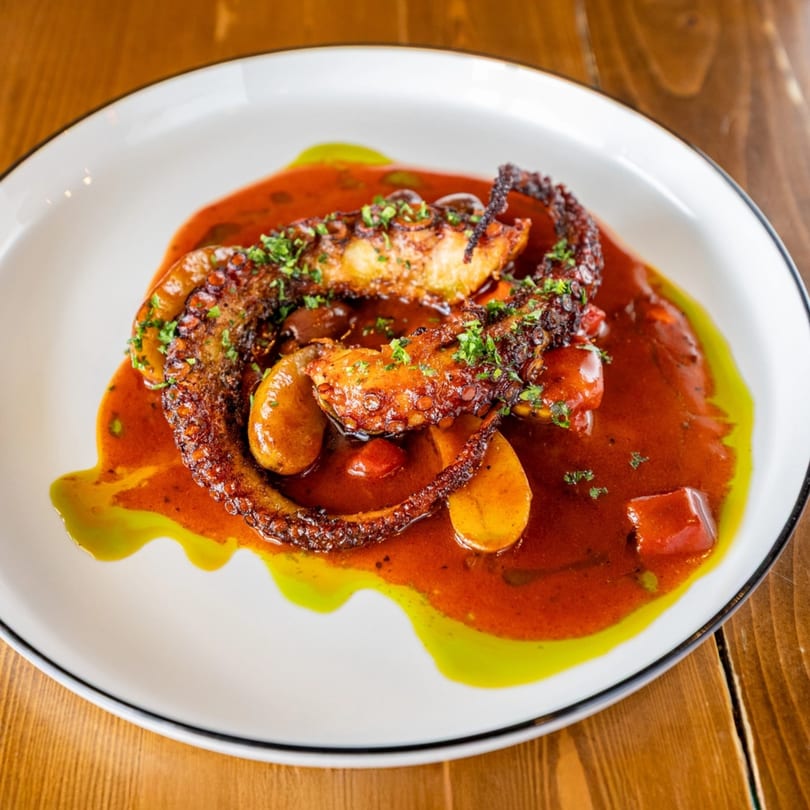 Grilled octopus tentacles with red sauce and green oil drizzle on a white plate.