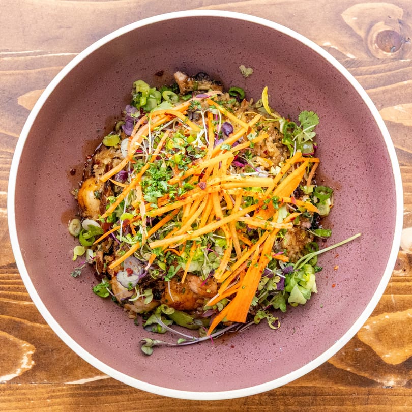 Caribbean dish in a purple bowl, topped with shredded carrots, herbs, and microgreens.
