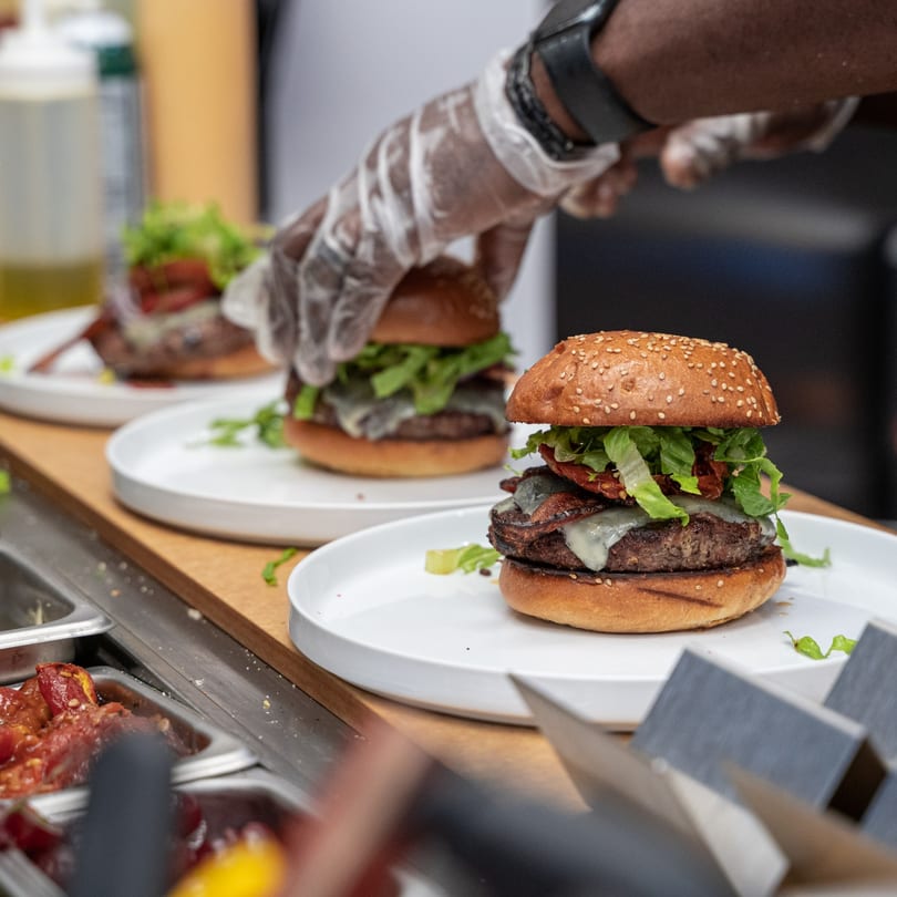 Gloved hands preparing multiple Caribbean-style burgers with cheese, bacon, and lettuce on white plates.