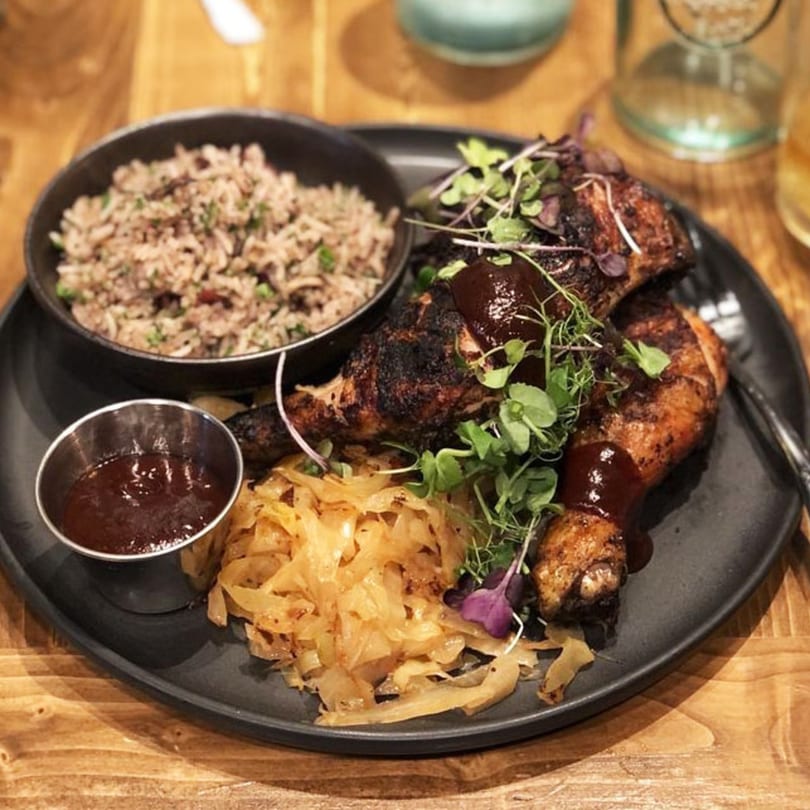 Caribbean jerk chicken with rice, sautéed cabbage, and barbecue sauce on a black plate.