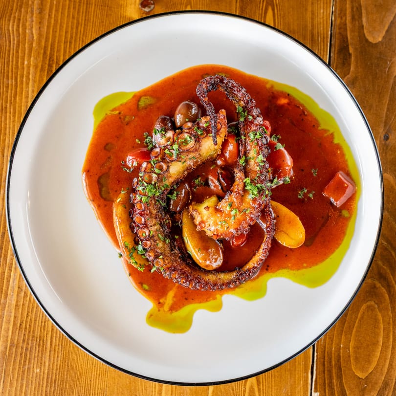Grilled octopus tentacles with potatoes, olives, and herbs in a rich red sauce with green oil.