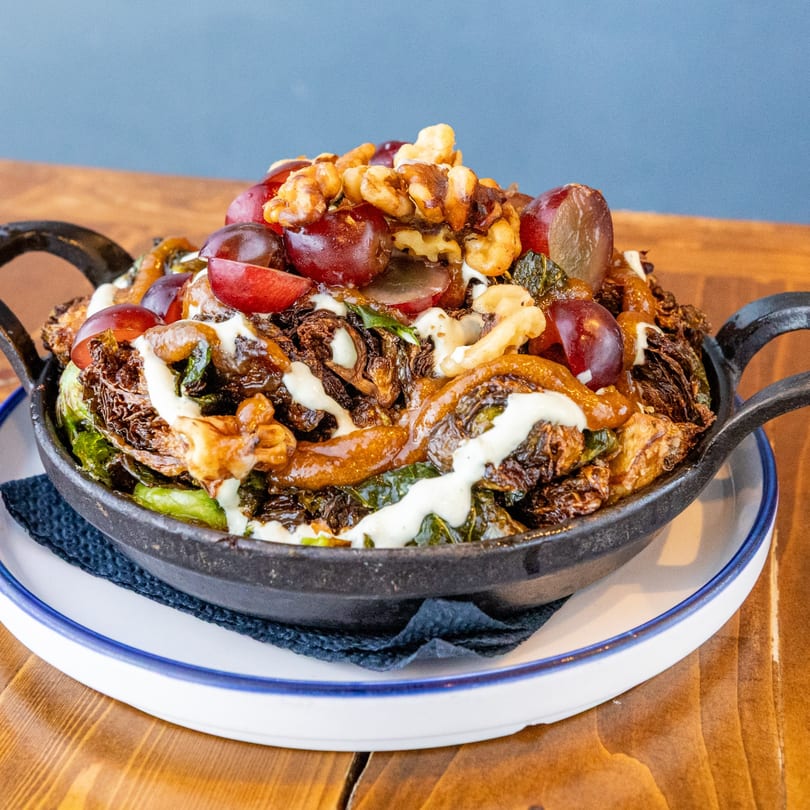 A Caribbean dish of roasted Brussels sprouts, grapes, walnuts, and white sauce in a cast iron pan.