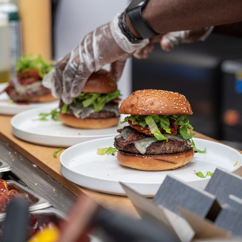 Gloved hands preparing multiple Caribbean-style burgers with cheese, bacon, and lettuce on sesame buns.