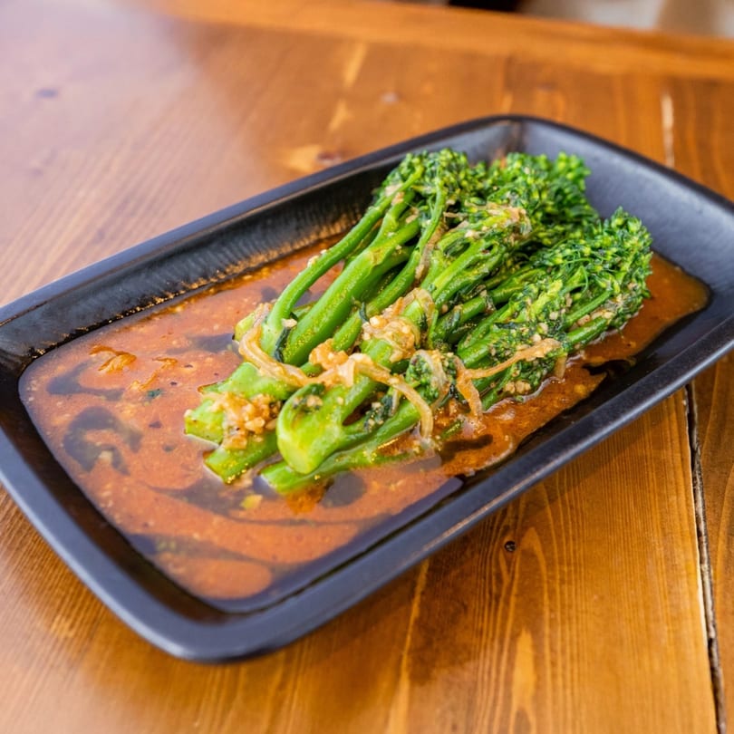 Broccolini with garlic and onions in a rich Caribbean sauce, served on a black rectangular plate.