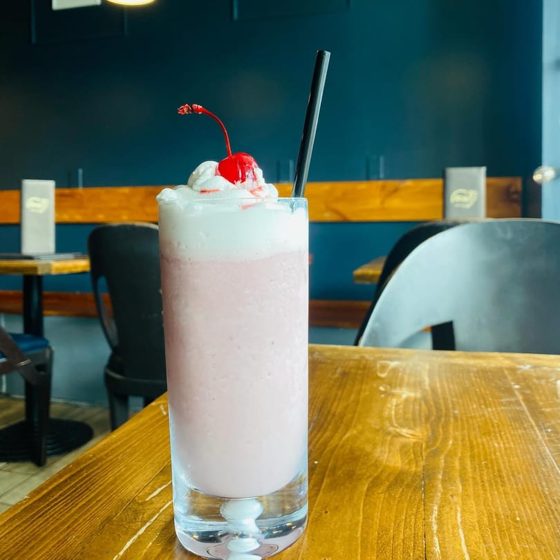 Pink Caribbean milkshake with whipped cream, a cherry, and a black straw on a wooden table.