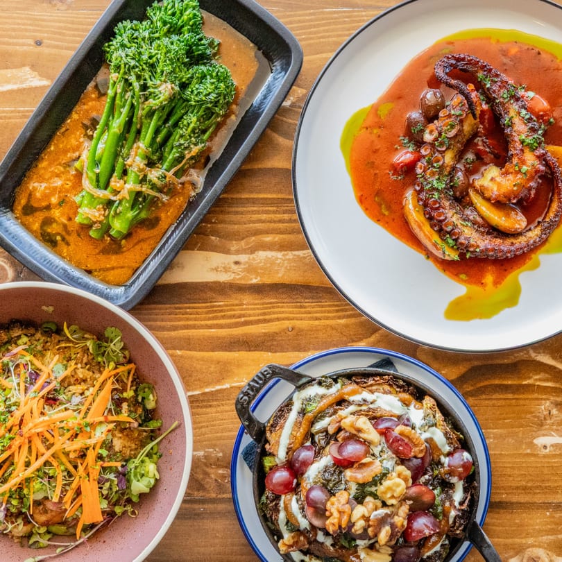 An overhead view of four Caribbean dishes: broccolini, octopus, salad, and a skillet dish.